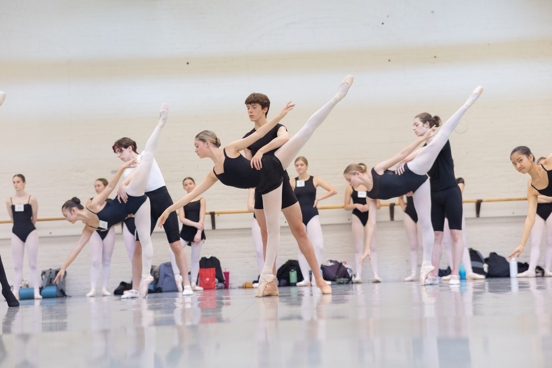 Male and female ballet dancers in rehearsal in a studio with large windows and bright light.