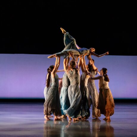 A group of BalletMet dancers stand in a circle and lift a single dancer above their heads.