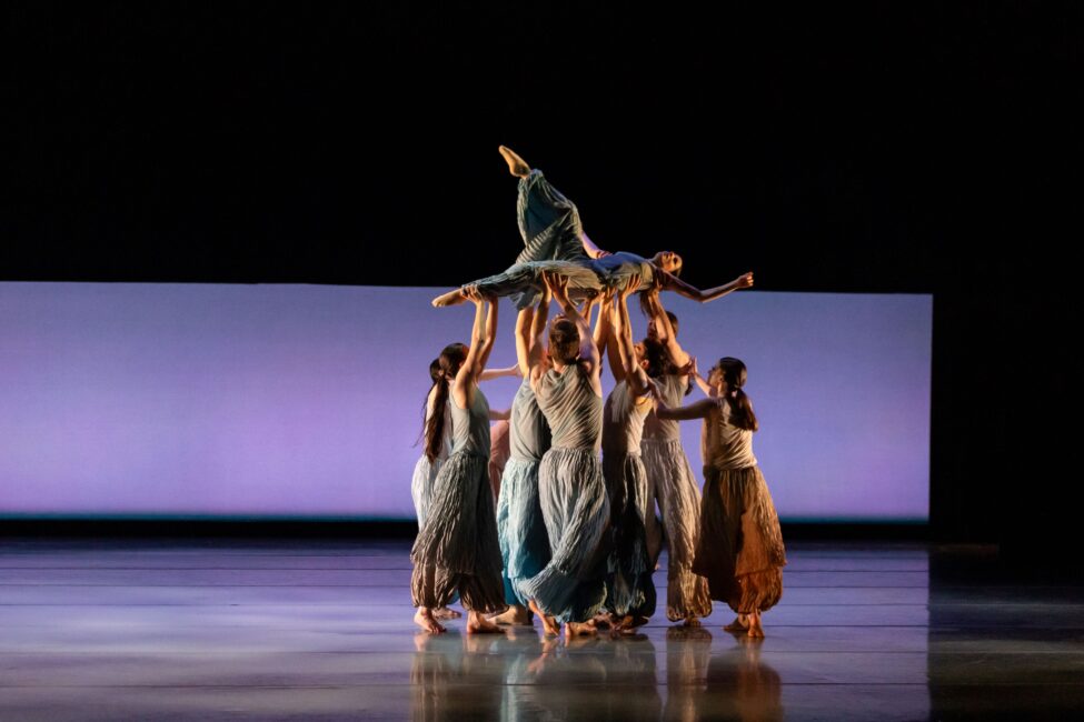 A group of BalletMet dancers stand in a circle and lift a single dancer above their heads.