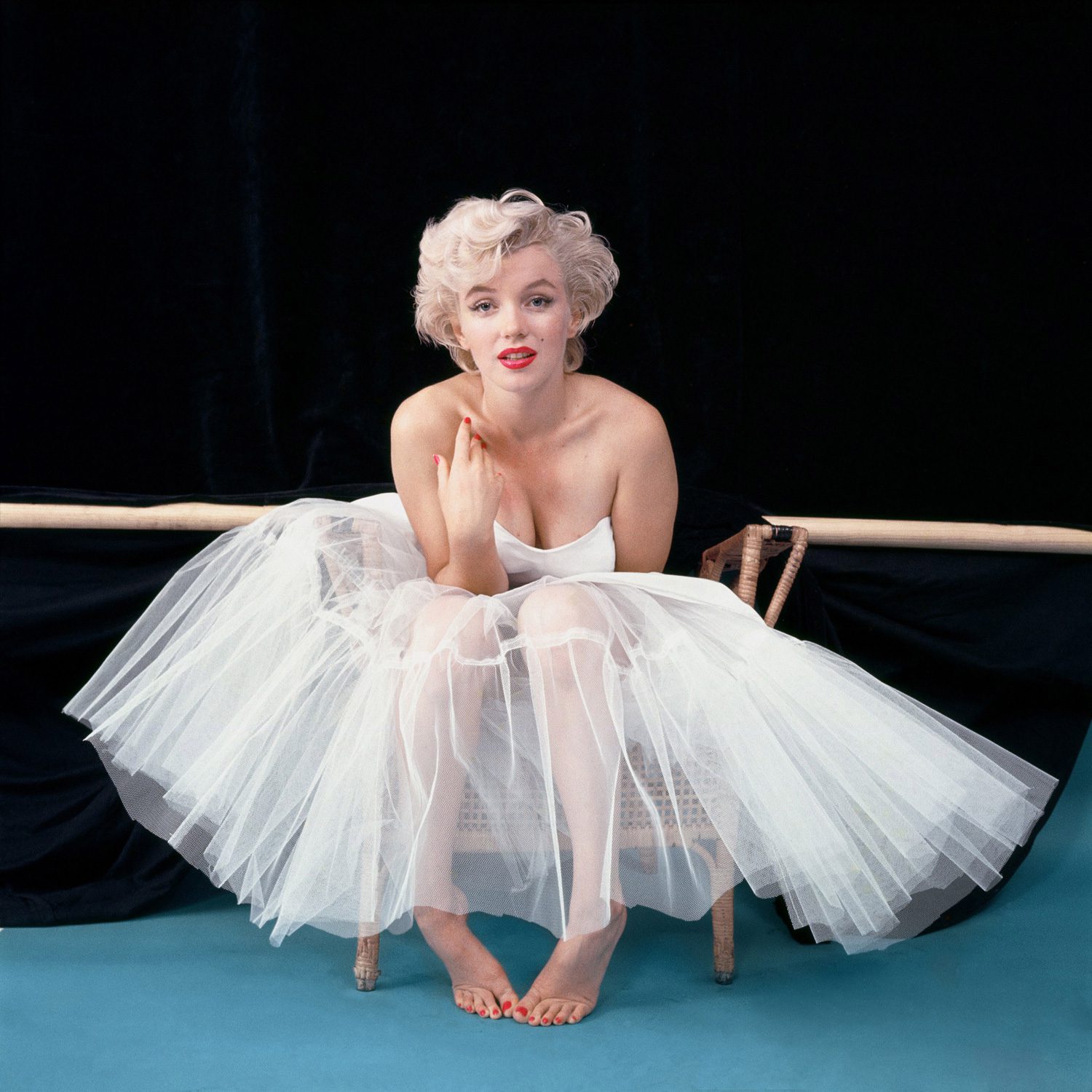 Marilyn Monroe with short blonde hair sits barefoot on a chair, wearing a white strapless tulle dress, evoking the classic elegance of her persona, against a black backdrop and blue floor.